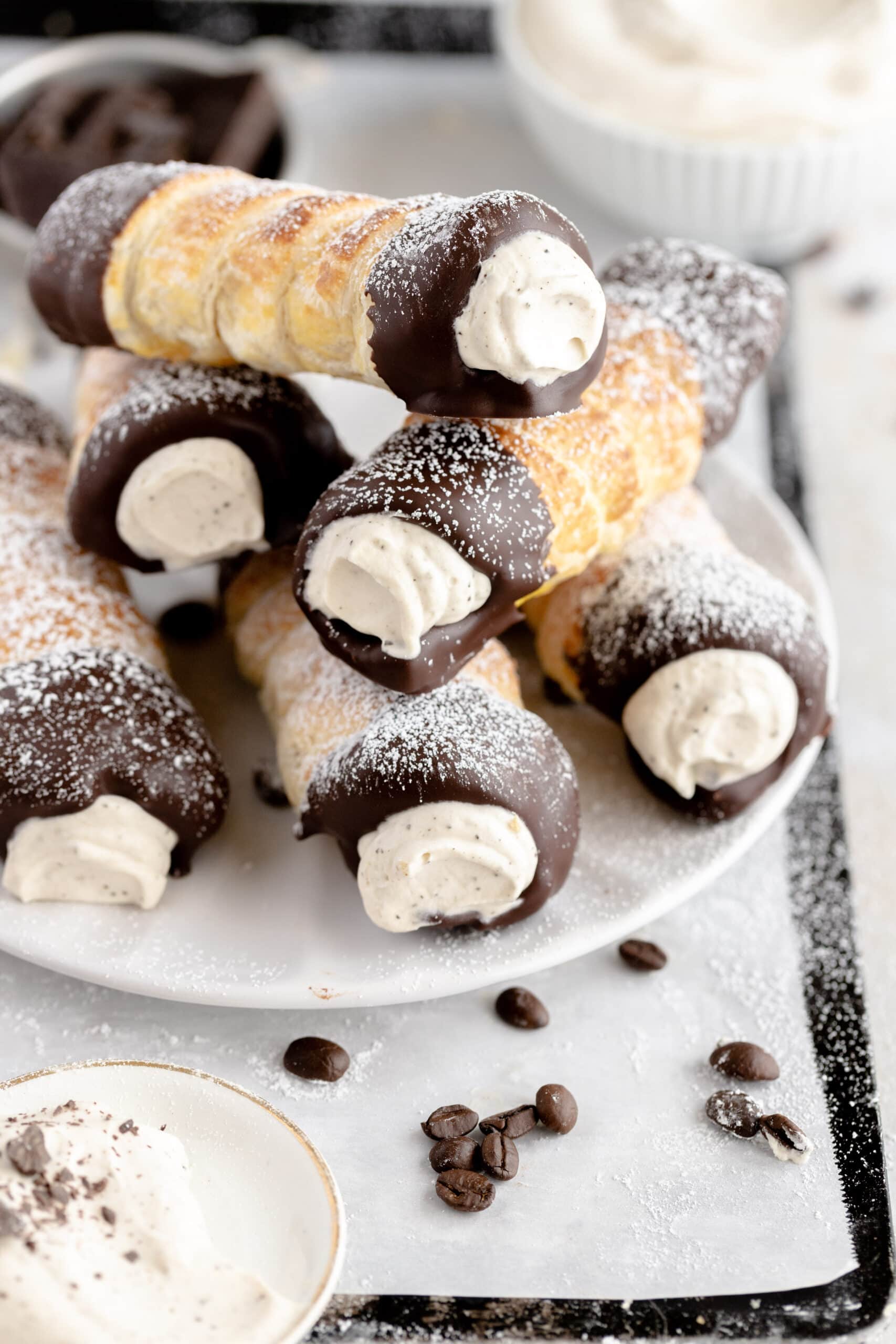Puff pastry rolls filled with coffee whipped cream and dipped in chocolate stacked on a white plate and dusted with powdered sugar.