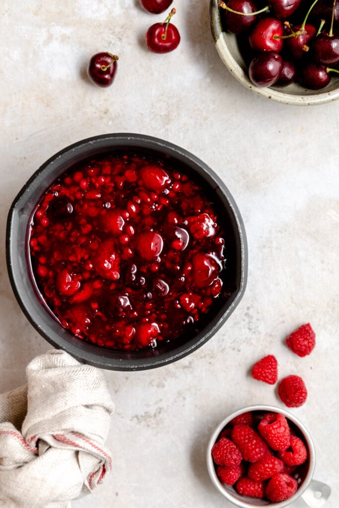German rote grütze in a black sauce pot surrounded by fresh raspberries and cherries.
