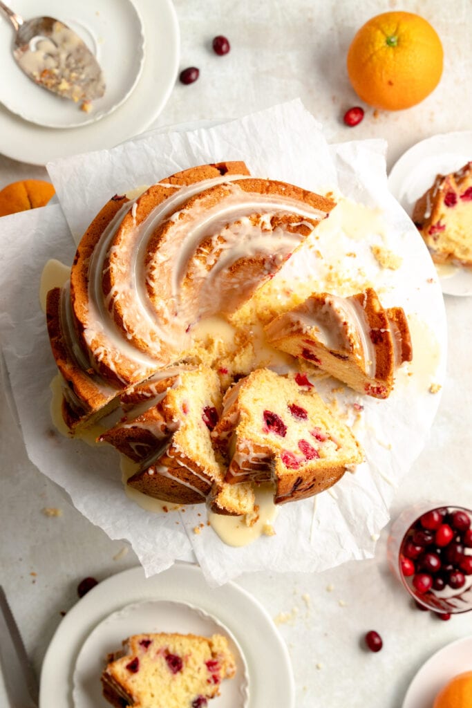 Overhead image of the Cranberry Orange Bundt Cake with orange glaze on a white cake stand with a few slices cut out of it.