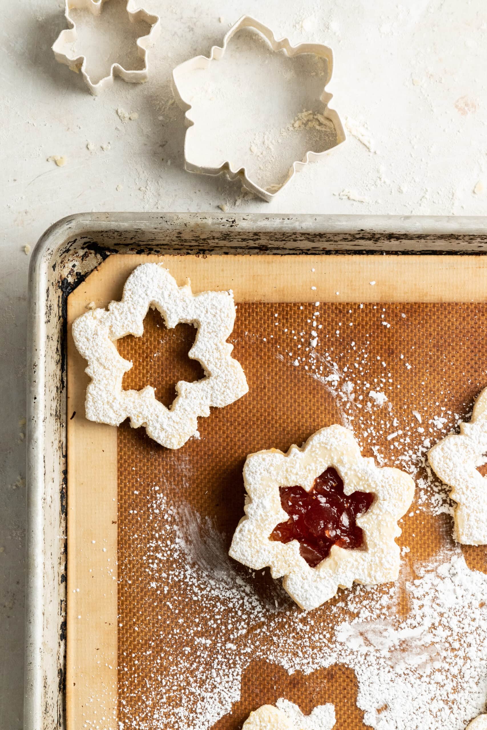 Overhead view of snowflake linzer plätzchen on a baking sheet with snowflake cookie cutters on the side.