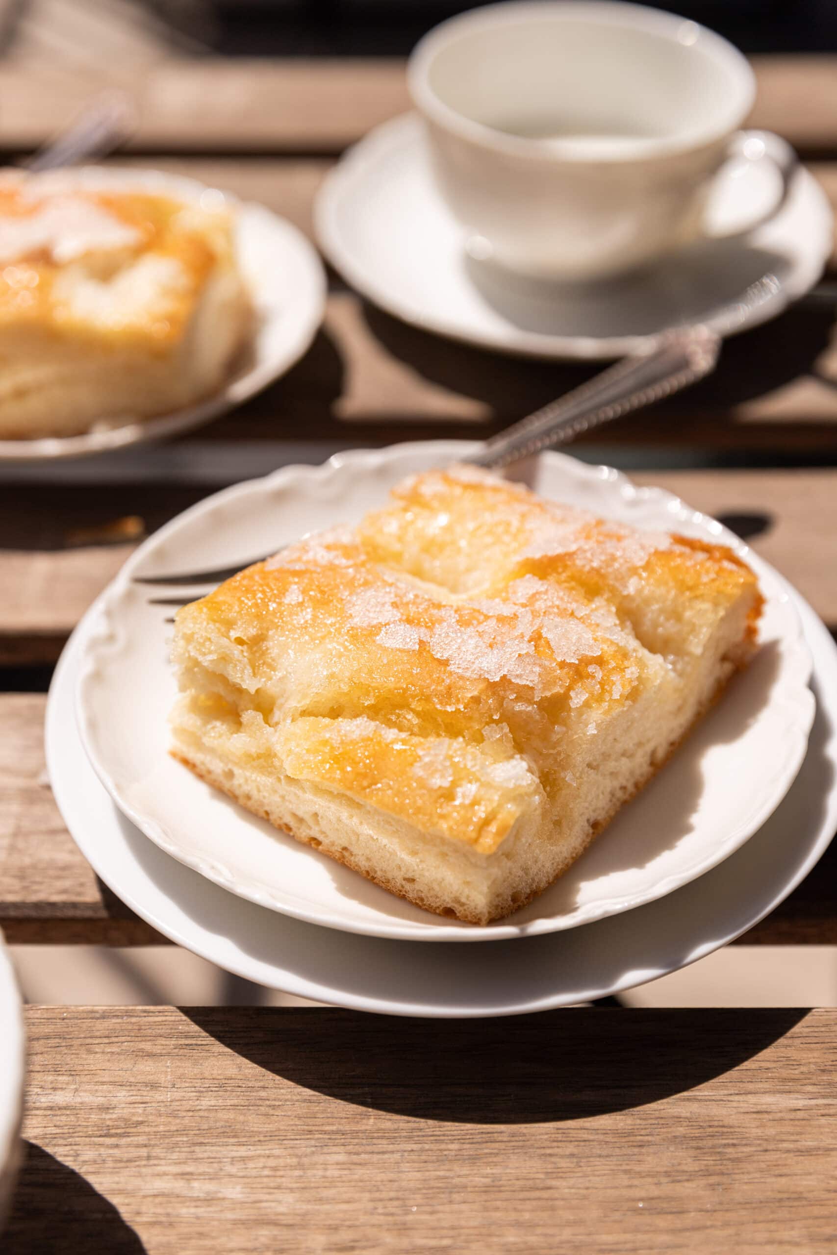 Side view of a square of butterkuchen on two white plates outside on a patio table with a coffee cup in the background. 