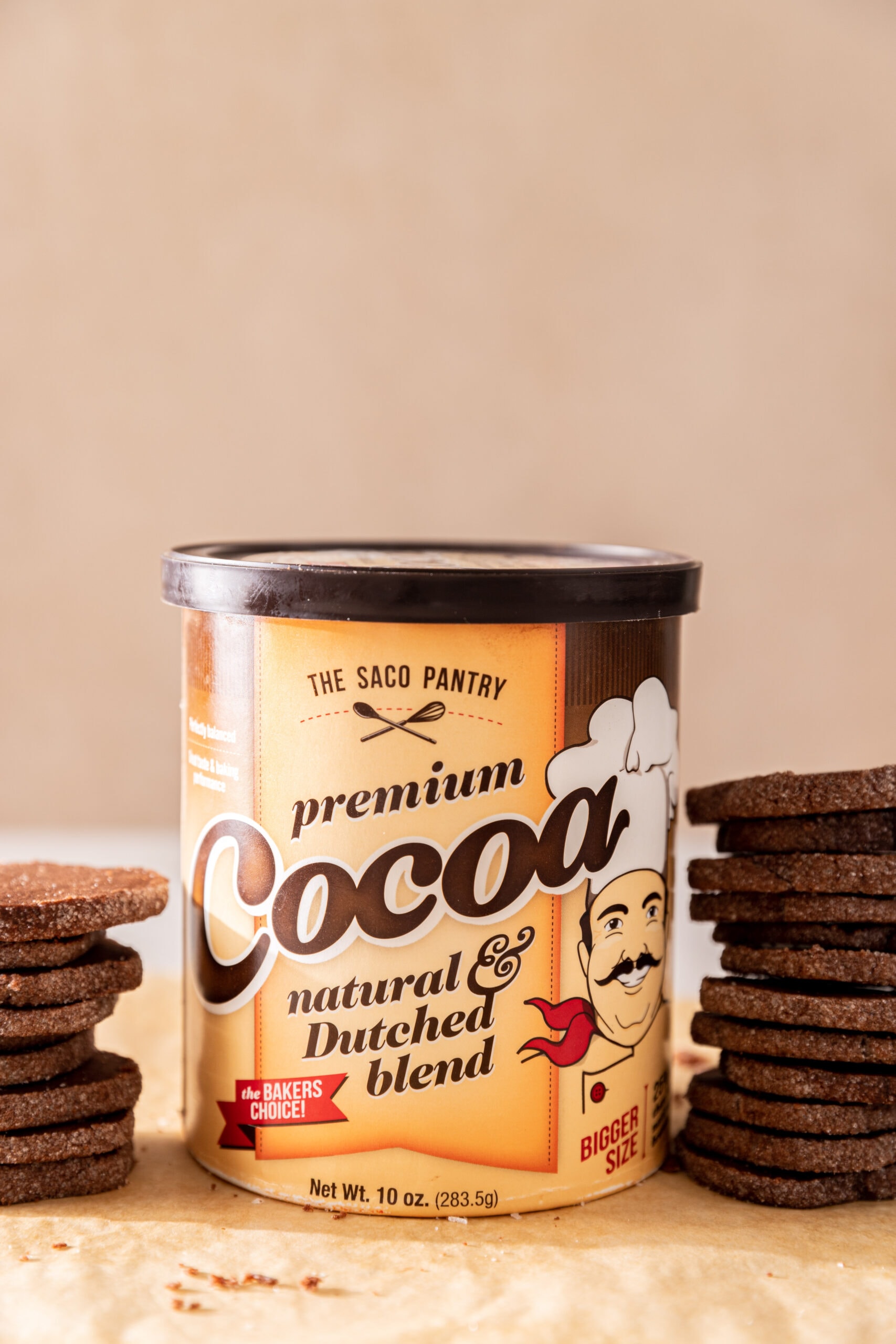 Large container of SACO Pantry Premium cocoa with two stacks of chocolate cookies.