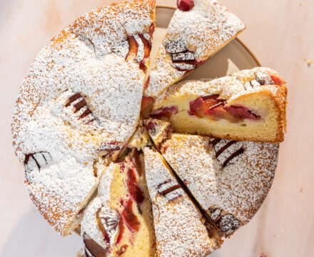 Overhead view of a plum cake, dusted with powdered sugar and cut into slices.