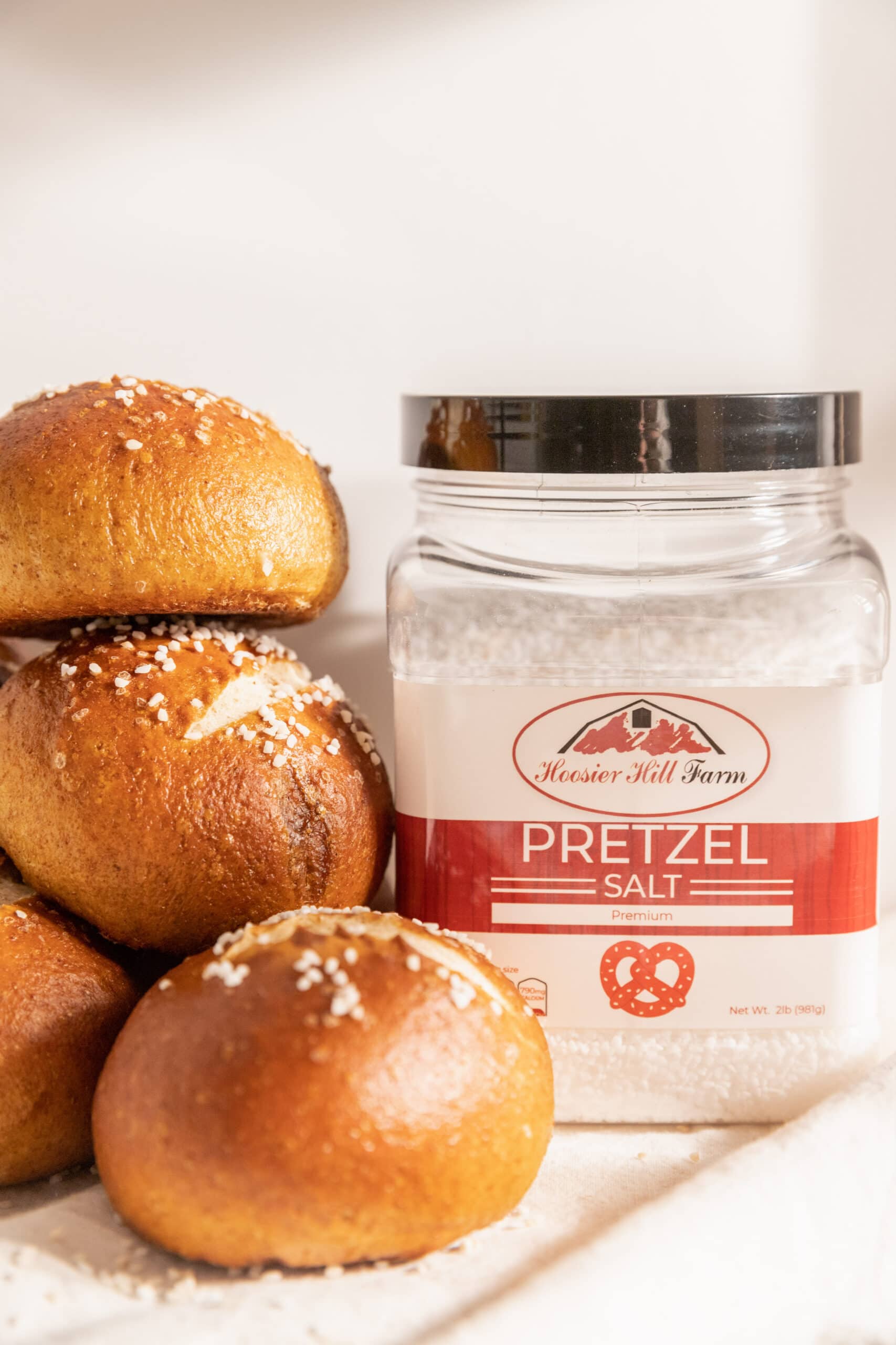 Side view of stacked pretzel rolls next to a container of Hoosier Hill Farm Pretzel Salt. 