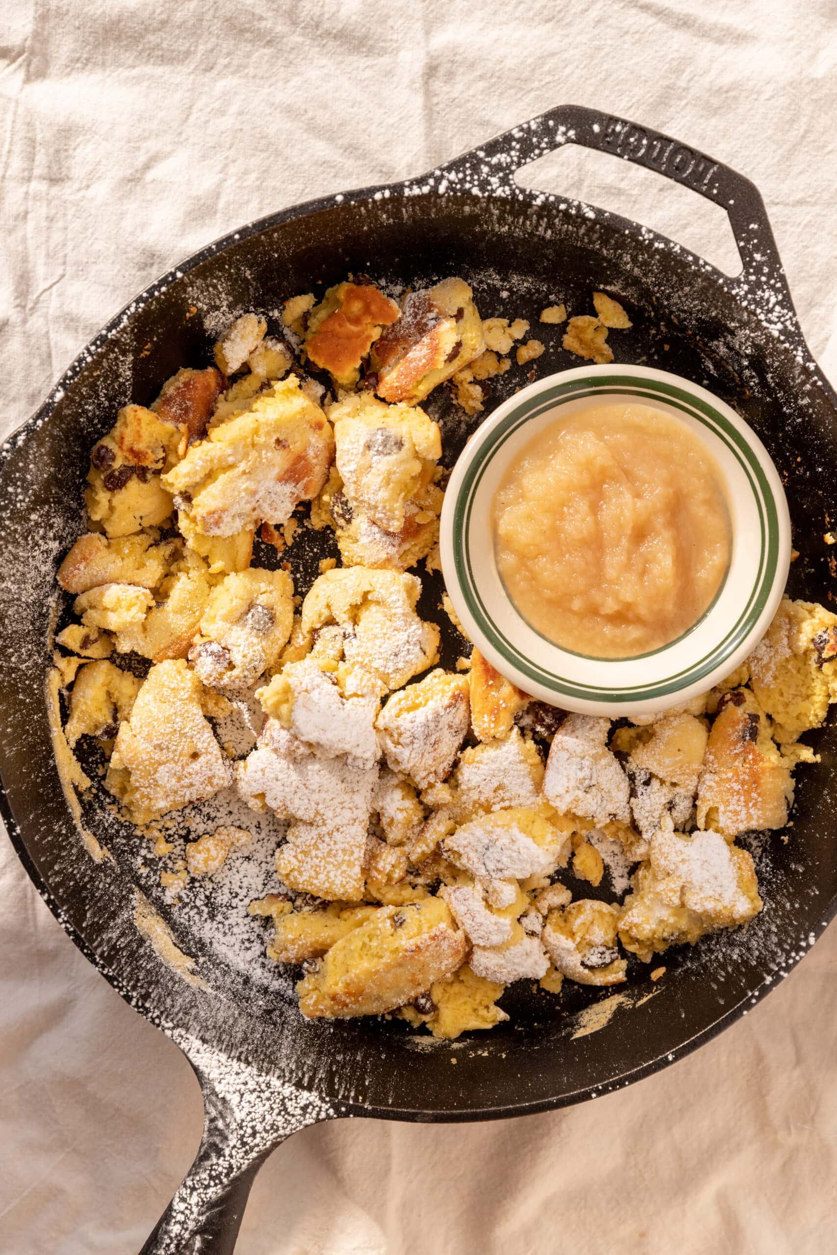 Overhead view of kaiserschmarrn in a cast iron pan, dusted with powdered sugar, and a bowl of apple sauce. 