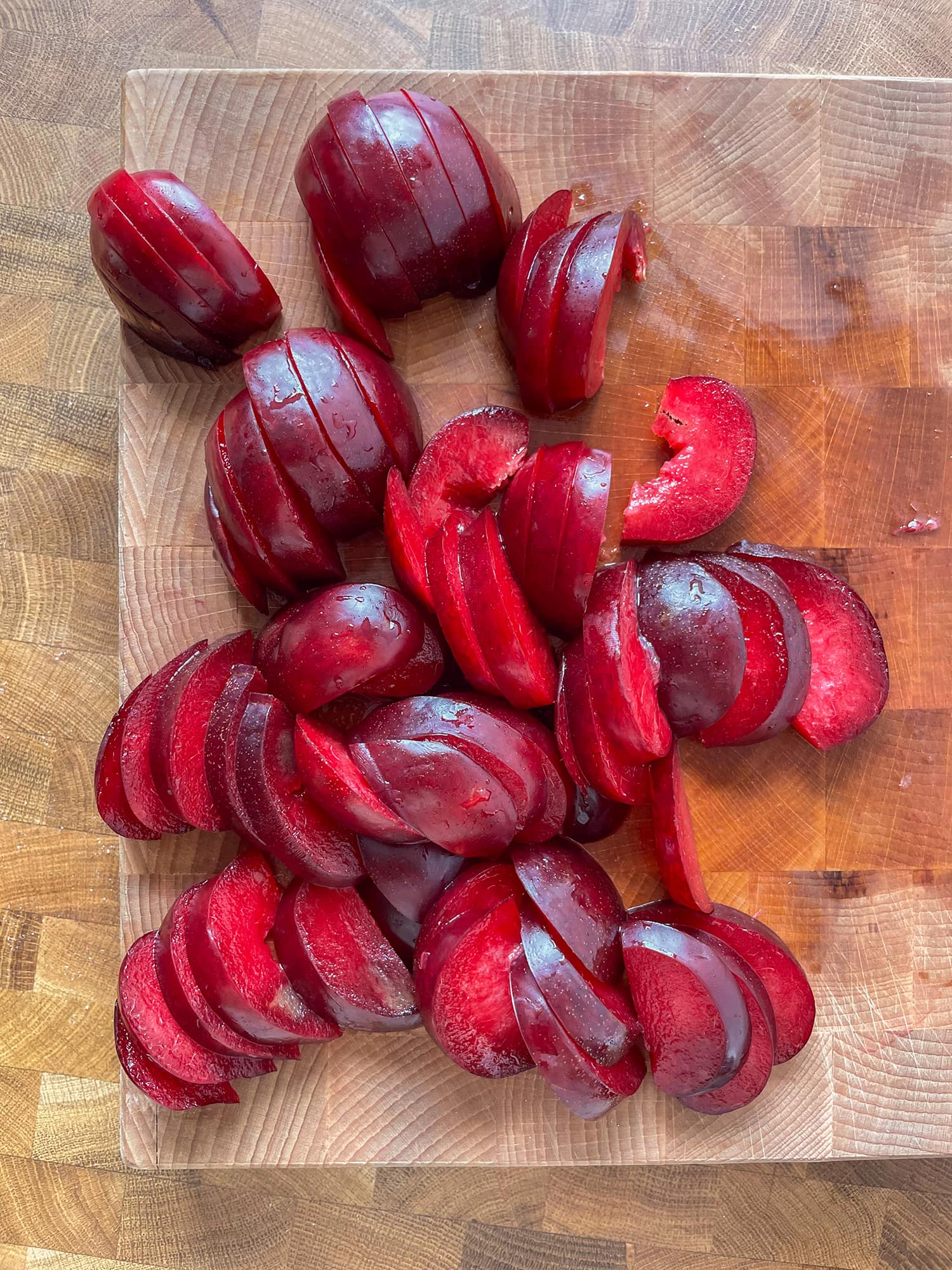 Red plums sliced into 1/2 inch slices on a wooden cutting board. 
