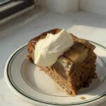 A slice of banana cake on a white and green plate topped with cream cheese whipped cream.