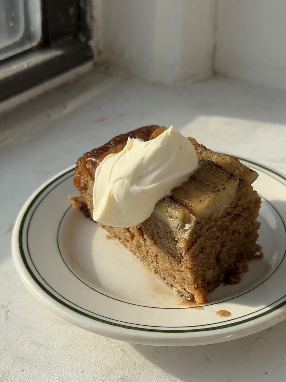 A slice of banana cake on a white and green plate topped with cream cheese whipped cream.
