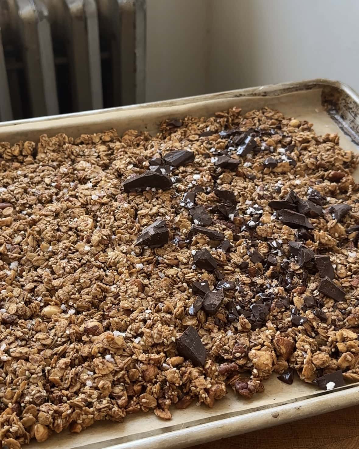 Side view of a sheet pan full of maple tahini granola, half topped with dark chocolate chunks.