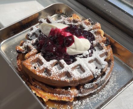 A stack of two blueberry waffles topped with blueberry compote, vanilla yogurt whipped cream, and a dusting of powdered sugar.