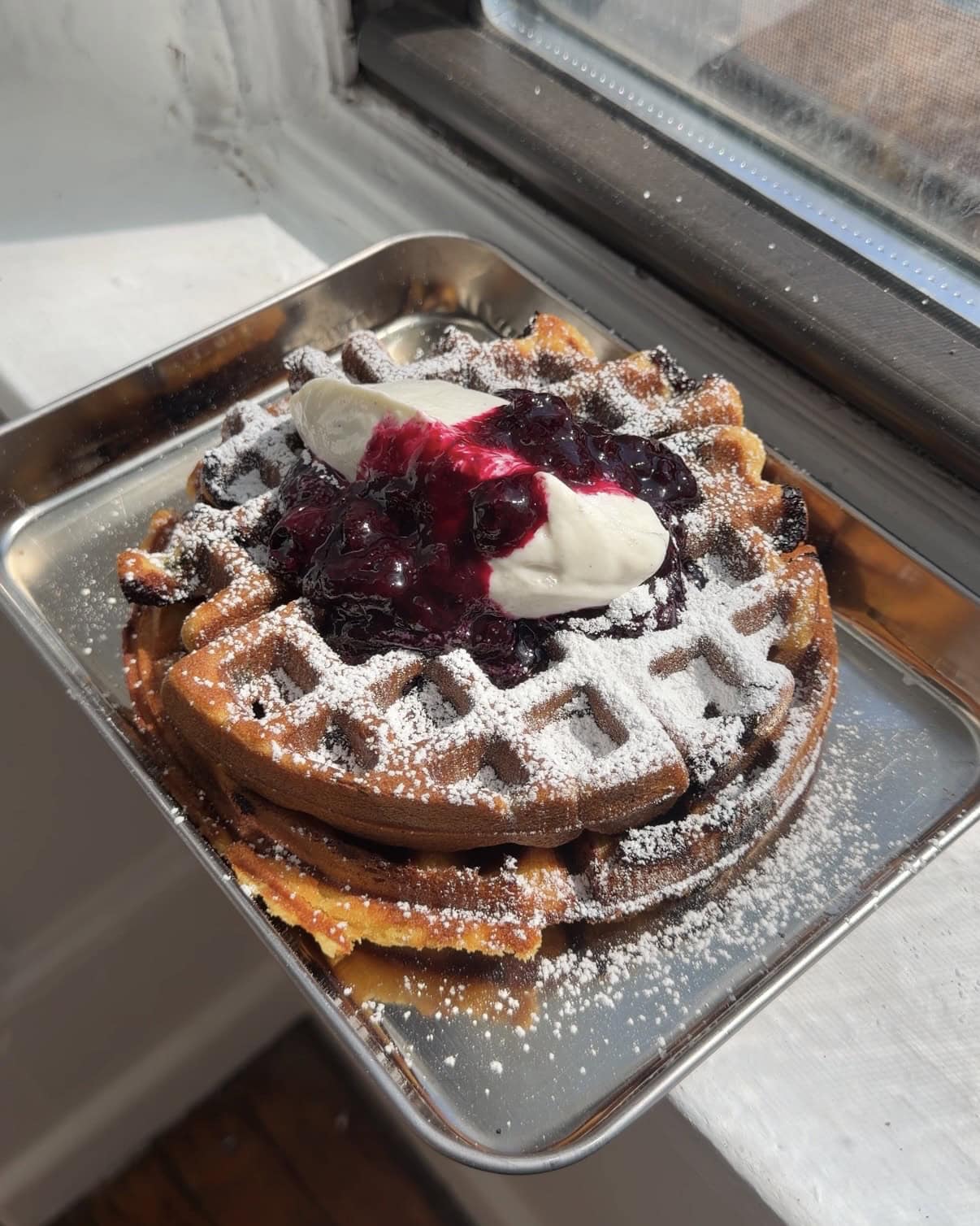 A stack of two blueberry waffles topped with blueberry compote, vanilla yogurt whipped cream, and a dusting of powdered sugar.
