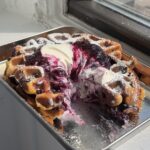 Side view of a stack of blueberry waffles with blueberry compote and yogurt whipped cream with bites taken out.