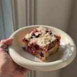 Side view of a slice of Plum Berry Crumb cake on a shallow white bowl in the sun.