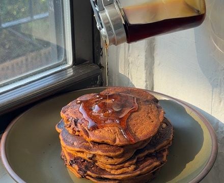 Side view of a stack of brown butter chocolate chip pumpkin pancakes being topped with maple syrup.